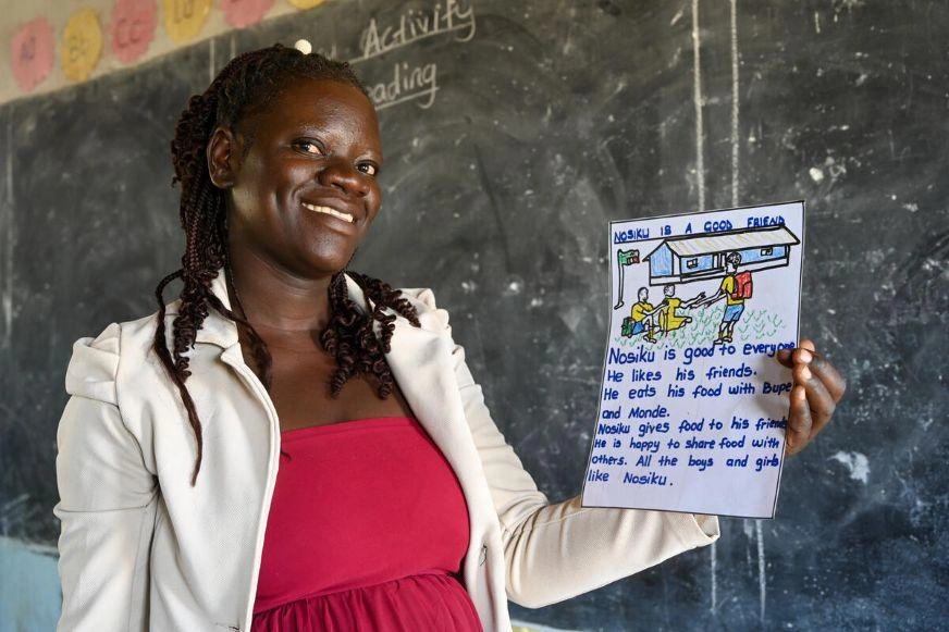 A lady smiles while holding up a hand-written story and hand-drawn picture of a boy named “Nosiku” who is a good friend to others.