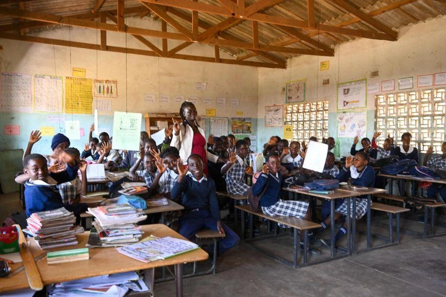 A classroom full of children wave toward the camera, big smiles on most faces. A woman stands amongst them.
