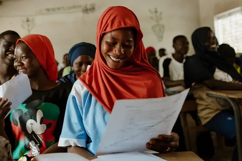 Young girl sitting in a classroom with other students, smiles while looking at an assignment on a sheet of paper.