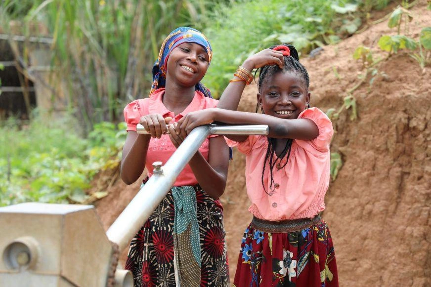 Two adolescent girls smile while leaning on a water pump. Both are wearing bright clothing.