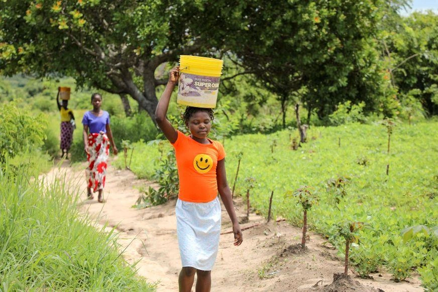 A girl carries a pail of water on her head. Two young women follow behind.